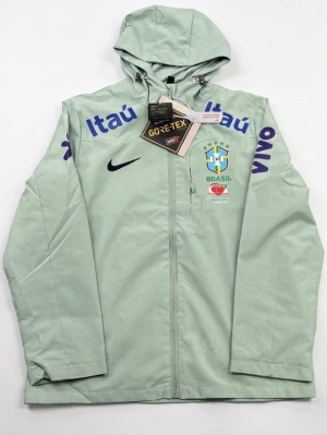 Nike Brasil vindjacka mintgrön - Mintgrön vindjacka från Nike med Brasil CBF-logga och sponsormärken som Itaú och Vivo. Jackan har huva, hel dragkedja och är tillverkad i GORE-TEX-material för extra skydd mot vind och regn. Perfekt för dig som gillar sportig stil och vill sticka ut.