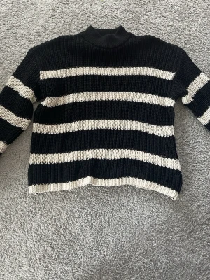 Svartvit randig stickad tröja - Säljer en stickad tröja med breda svarta och vita ränder. Tröjan har hög hals och långa ärmar, perfekt för kyligare dagar. Grovstickad och mjuk, passar till jeans eller kjol för en avslappnad look. Aldrig använd.