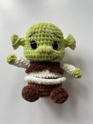 Virkad Shrek-inspirerad mjukis - Säljer en handgjord, stickad mjukis som liknar Shrek. Dockan är grön med stora svarta ögon, bruna och vita kläder samt mjuka, fluffiga material. Perfekt som dekoration eller present till någon som gillar animerade filmer. 18,5 cm hög