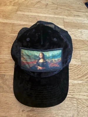 Svart keps med Mona Lisa-tryck - Snygg svart keps med mesh baktill och ett stort tryck av Mona Lisa på framsidan. Kepsen har böjd skärm och justerbar rem bak. Materialet är en mix av tyg och mesh som ger en cool och luftig känsla.