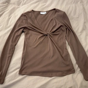 Brun omlottopp från Vila - Säljer en brun långärmad topp från Vila med omlottdetalj och twist framtill. Toppen har en djup v-ringning och är tillverkad i ett mjukt, stretchigt material som sitter snyggt på kroppen. Perfekt att styla med jeans eller kjol.
