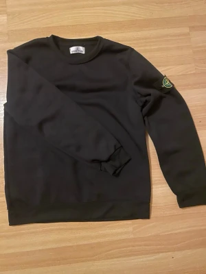 Stone Island sweatshirt  - Svart sweatshirt från Stone Island med klassisk rund hals och ikonisk logotyp-patch på vänster ärm. Tröjan har en stilren design med ribbade muddar vid ärmslut och nederkant. Perfekt för en avslappnad och trendig look. 