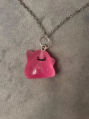 Självlysande Ditto halsband Pokémon smycke  - Så gulligt halsband med en berlock av Ditto!Berlocken är ca 1,5 x 1,5 cm och trädd på en kedja i silverimitation, justerbar 40-45 cm. Betlocken är också självlysande och lyser i mörkret.