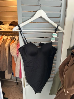 Svart shapingbody  - Snygg svart shapingbody från DORINA med vadderade kupor och justerbara axelband. Bodyn har en slät och stretchig design som formar kroppen och ger stöd. Perfekt under tighta plagg för en smickrande siluett. Materialet är mjukt och följsamt.