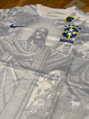 Vit Brasilien fotbollströja Nike - Snygg vit Brasilien fotbollströja från Nike med tryck av Kristusstatyn och detaljer i blått och gult. Tröjan har rund hals, korta ärmar och CBF-emblem på bröstet. Perfekt för dig som älskar fotboll och vill sticka ut med en unik design.