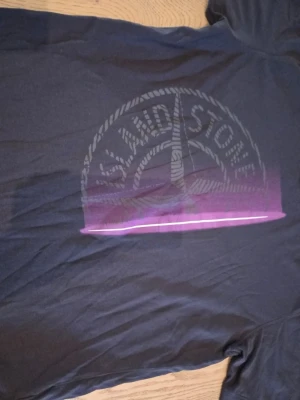 Stone Island t-shirt - Snygg mörkgrå t-shirt från Stone Island med stort tryck på bröstet i grått och lila. Klassisk rund halsringning och korta ärmar. Perfekt för dig som gillar streetwear och vill sticka ut med en ikonisk logga.