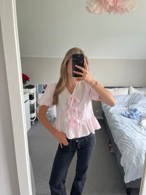 Söt rosa blus med rosetter - Ljusrosa blus med korta puffärmar och volang nertill. Blusen har tre dekorativa rosetter framtill och v-ringning. Tillverkad i ett lätt och luftigt tyg. Andvänd fåtal gånger💕 Pris går att diskutera 