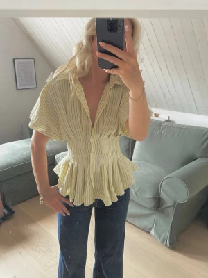 Gulrandig blus med volang - Trendig gul blus med tunna svarta ränder och volang nedtill. Blusen har korta ärmar, markerad midja och knappar framtill. Perfekt för dig som gillar en feminin och chic stil. Materialet ser ut att vara lätt och luftigt, vilket gör den skön att bära.
