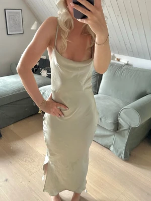 Ljusgrön långklänning med öppen rygg - Säljer en ljusgrön långklänning i satinliknande material med smala axelband och snygg öppen rygg med snörning. Klänningen har en slits i sidan och en elegant, figurnära passform. Perfekt för dig som vill ha en stilren och trendig look. OBS, har en liten vattenfläck som man ser på första bilden som säkert är enkel att få bort 