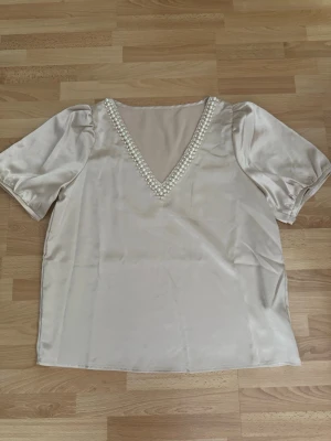 Ny.Champagnefärgad blus med pärldetaljer - Snygg champagnefärgad blus i glansigt satintyg/polyester med korta puffärmar och djup V-ringning dekorerad med pärlor. Perfekt för dig som vill ha en elegant och trendig look. Materialet känns lyxigt och skimrande. Bröst 110cm, längd 63cm.