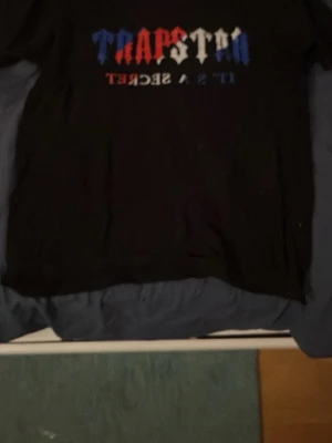 Trapstar svart t-shirt med tryck - Säljer en svart t-shirt från Trapstar med stort tryck i rött, vitt och blått på bröstet där det står 'Trapstar It's a secret'. Klassisk passform och korta ärmar. Perfekt för dig som gillar streetwear och vill sticka ut.