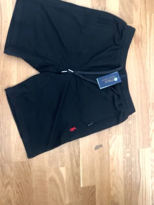 Svarta shorts från Polo Ralph Lauren - Svarta shorts från Polo Ralph Lauren med klassisk passform och elastisk midja med snörning. Liten röd logga broderad framtill. Tillverkade i mjuk bomull, perfekta för en avslappnad stil.