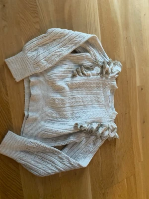 Vit stickad tröja med volangdetalj - Supersöt vit stickad tröja med långa ärmar och volangdetaljer med guldtråd på axlarna. Tröjan har ett klassiskt kabelstickat mönster och ribbade muddar. Perfekt för dig som vill ha något extra till din outfit.