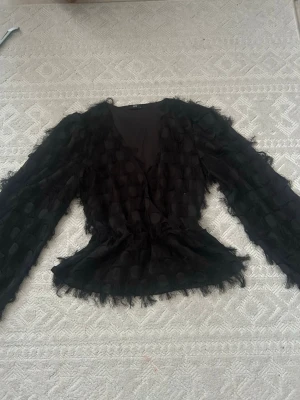 Svart meshblus med prickar från JDY - Trendig svart meshblus från JDY med långa ärmar och markerad midja. Blusen har ett coolt prickmönster i sammet och fransiga detaljer som ger en unik look. Tunn och lätt material, perfekt för att styla lager på lager. Djup v-ringning framtill.