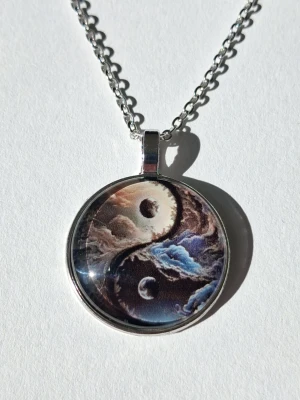 Yin Yang halsband med galaxmotiv - Unikt halsband med ett runt hängsmycke i metall och 2D som har ett färgstarkt yin yang-motiv med galax och moln i blått, vitt och svart. Kedjan är silverfärgad och länkarna är medelgrova. Materialet rostfritt stål 