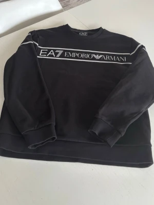 Svart sweatshirt från EA7 Emporio Armani - Svart sweatshirt från EA7 Emporio Armani med vit logga och text framtill. Tröjan har rund halsringning, ribbade muddar vid ärmslut och nederkant samt en klassisk, avslappnad passform. Perfekt för en trendig och sportig look.