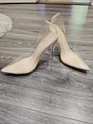 Transparenta pumps med spetsig tå - Snygga pumps med transparent ovandel och spetsig tå. Skorna har en beige innersula och höga klackar i klar plast. Perfekta för dig som vill ha en modern och trendig look. Klackarna är smala och ger en elegant siluett.