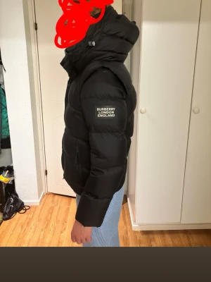 Svart dunjacka från Burberry - Snygg svart dunjacka från Burberry med huva och tryckt logga på ärmen. Jackan har en puffig design, dragkedja och knappar framtill samt praktiska sidofickor. Perfekt för kalla dagar och ger en stilren look.