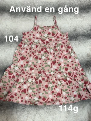 Rosa jordgubbsklänning med volang - Söt rosa klänning med jordgubbsmönster och vita blommor. Klänningen har smala axelband och volang i kjolen, perfekt för varma dagar. 🌸 Använd en gång. Storlek 104
