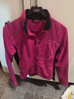 Rosa vindjacka från Everest - Snygg rosa vindjacka från Everest med svarta detaljer på sidorna och insidan av kragen. Jackan har dragkedja framtill, en bröstficka med dragkedja och reflekterande detaljer. Perfekt för blåsiga dagar och outdoor-aktiviteter.