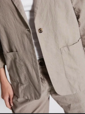 Beige kavaj från 157 - Stilren beige kavaj från 157 med avslappnad passform och stora utanpåfickor. Kavajen har enkelknäppning med en knapp och är tillverkad i ett lätt, skrynkligt tyg som ger en cool och modern look. Perfekt för dig som gillar minimalistisk stil.