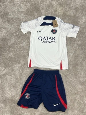 PSG fotbollskit Nike vit/blå - Snyggt PSG fotbollskit med vit tröja och blå shorts. Tröjan har PSG-logga, Qatar Airways-tryck och röda detaljer. Shortsen är mörkblå med röda ränder och PSG-märke. Perfekt för fotbollsträning eller match. Materialet är lätt och andas.