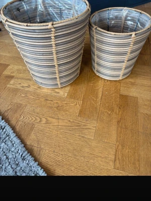 Två flätade korgar i rotting - Säljer två snygga korgar i naturfärgad rotting med plastad insida. Perfekta som blomkrukor eller förvaring. Korgarna har en rund form och stilrena ränder i beige och grått. Passar bra i hemmet för att skapa en mysig känsla.