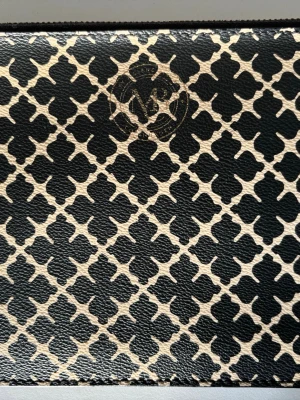 Svart och beige mönstrad clutch - Säljer en clutch från By Malene Birger i svart och beige med deras ikoniska blommönster. Väskan är tillverkad i konstläder och har en diskret logga framtill. Perfekt för att bära småsaker eller använda som necessär.