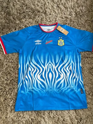 Blå Kongo DR matchtröja Umbro XL - Säljer en blå fotbollströja från Umbro för Kongo DR med zebramönster, röda och vita detaljer vid krage och ärmslut. Tröjan har FECOFA-emblem, flagga och två stjärnor på bröstet. Perfekt för dig som älskar unika matchtröjor!