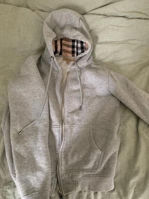 Grå hoodie från Burberry med dragkedja - Snygg grå hoodie från Burberry med klassiskt rutigt foder i huvan. Dragkedja framtill, två fickor och justerbara snören i huvan. Mjuk insida och ribbade muddar. Perfekt för en stilren och avslappnad look.