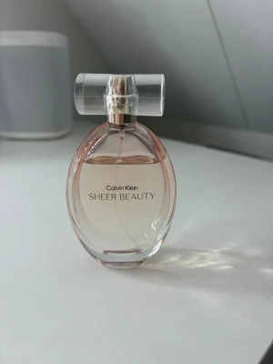 Calvin Klein Sheer Beauty parfym - Elegant parfymflaska från Calvin Klein med namnet Sheer Beauty. Flaskan är oval och transparent med en svagt rosa ton och har ett stilrent, genomskinligt lock. Perfekt för dig som gillar fräscha och feminina dofter.