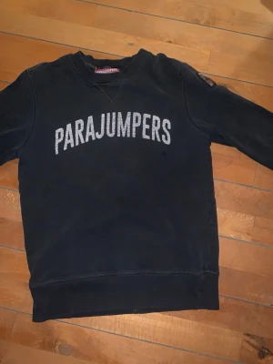 Svart sweatshirt från Parajumpers - Cool svart sweatshirt från Parajumpers med stort vitt tryck över bröstet. Klassisk rund halsringning och ribbade muddar vid ärmslut och nederkant. Perfekt för en avslappnad streetstyle-look.