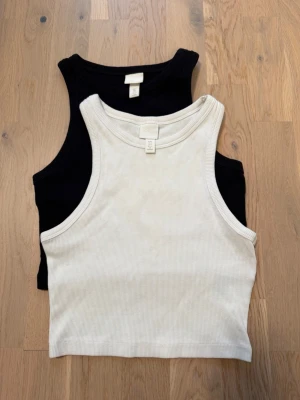 2-pack ribbade linnetoppar H&M - Två ribbade linnetoppar från H&M, en i svart och en i vit. Båda är ärmlösa med rund halsringning och croppad passform. Perfekta att matcha med jeans eller kjol för en clean och enkel look 🤍