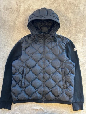 Moncler cardigan - Snygg svart pufferjacka från Moncler med quiltat mönster, huva och dragkedja framtill. Jackan har svarta ärmar i mjukare material och Moncler-logga på vänster ärm. Perfekt för kalla dagar och riktigt trendig look. Storlek xxl passar xl behöver ni fler bilder så hör av er bara 