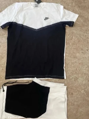 Nike svartvitt set med t-shirt och shorts - Snyggt svartvitt set från Nike med t-shirt och matchande shorts. T-shirten har en vit överdel och svart nedtill med Nike-logga på bröstet. Shortsen är vita med svart panel och dragsko i midjan. Perfekt för en sportig och avslappnad stil.