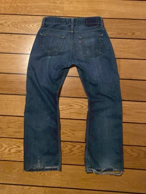 Blå raka jeans från Levi's - Säljer ett par klassiska blå Levi's jeans med rak passform och slitningar på vänster lår samt vid bensluten. Jeansen har fem fickor, knappgylf och är tillverkade i kraftig denim. Perfekta för en avslappnad och trendig look.
