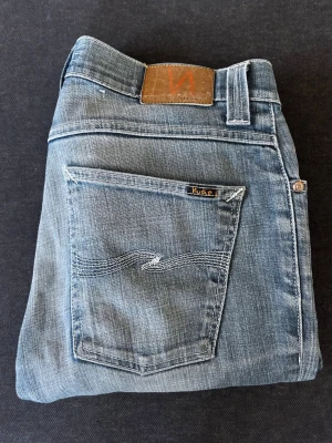 Grim tim jeans från Nudie - Snygga blå slim fit jeans från Nudie med klassisk femficksdesign och diskreta slitningar. Jeansen har en ljus tvätt och ikonisk vågstickning på bakfickan. Perfekta för en avslappnad och trendig stil. Modellen är Grim Tim 32/32!