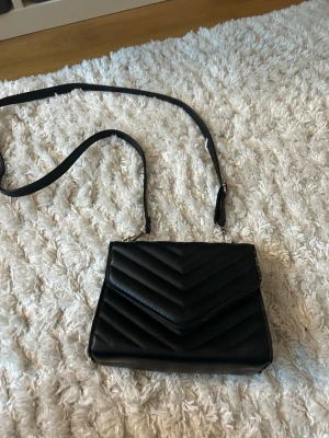 Svart quiltad axelväska i skinnimitation - Snygg svart axelväska med quiltat mönster i chevron-stil. Väskan har en justerbar axelrem och stängs med lock. Tillverkad i skinnimitation och har en stilren, rektangulär form som passar till många outfits.