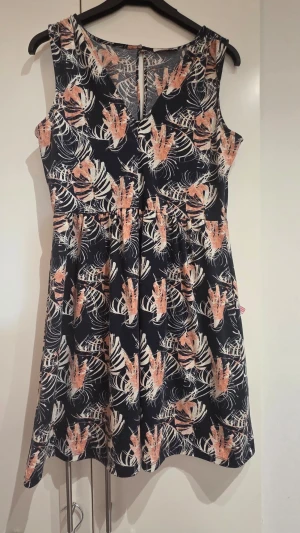 Mönstrad ärmlös kortklänning - Snygg ärmlös kortklänning med marinblå botten och tropiskt mönster i beige och orange. Klänningen har en markerad midja och rund halsringning. Perfekt för varma dagar och ger en fräsch sommarkänsla.