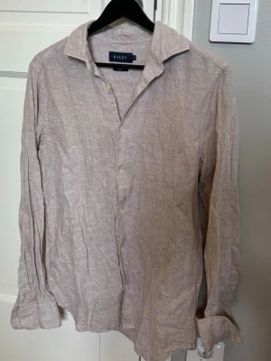 Beige linneskjorta från Riley - Säljer en stilren beige skjorta från Riley i 100% linne. Skjortan har klassisk krage, knäppning framtill och långa ärmar. Perfekt för en avslappnad men snygg look. Bara att höra av er vid frågor eller funderingar!