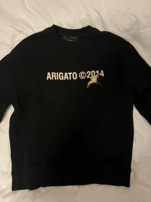 Svart sweatshirt Axel Arigato - Svart sweatshirt från Axel Arigato med trycket 'ARIGATO ©2014' och en liten broderad fågel på bröstet. Klassisk rund halsringning och ribbade muddar. Perfekt för en avslappnad streetwear-look.