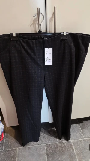 Mörka byxor med diskret rutmönster - Snygga mörka byxor från Robell  i storlek 48. Byxorna har ett diskret rutmönster och rak passform. Perfekta för dig som vill ha en stilren look med lite extra detalj. Materialet känns mjukt och bekvämt, troligen polyesterblandning.