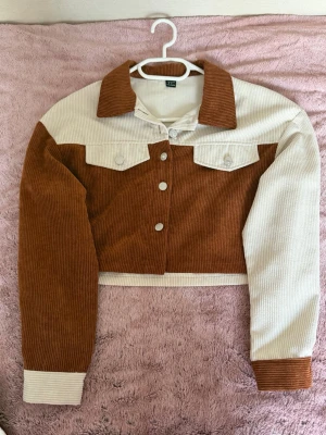Tvåfärgad manchesterbyxdress - Säljer en unik byxdress i manchestertyg med cool tvåfärgad design i beige och rostbrunt. Jackan är croppad med bröstfickor och matchande knappar, byxorna har raka ben och hög midja. Perfekt för dig som vill sticka ut med retrovibbar!