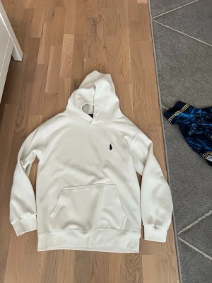 Vit hoodie från Polo Ralph Lauren - Vit hoodie från Polo Ralph Lauren med klassisk svart broderad logga på bröstet. Hoodien har känguruficka framtill, ribbade muddar och är tillverkad i mjukt material. Perfekt för en clean och stilren look.