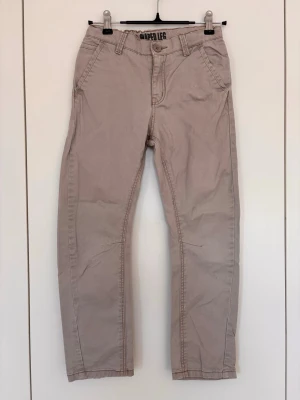 Beige chinos, HM, strlk 128 - Snygga beige chinos från Lappeljeans med raka ben och klassiska fickor fram och bak. Byxorna har bälteshällor, knappgylf och är tillverkade i ett mjukt bomullstyg som ger en avslappnad look. Perfekta för dig som gillar stilrena och enkla byxor.