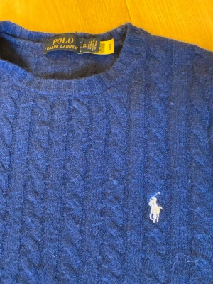 Mörkblå kabelstickad tröja Polo Ralph Lauren - Kabelstickad mörkblå tröja från Polo Ralph Lauren med rund halsringning och klassisk broderad logga på bröstet. Tröjan har långa ärmar och ribbade muddar vid ärmslut och nederkant. Perfekt för en stilren och tidlös look.