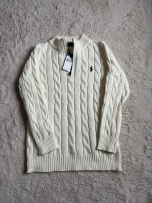 Vit kabelstickad tröja från Polo Ralph Lauren - Kabelstickad vit tröja från Polo Ralph Lauren med klassisk svart logga broderad på bröstet. Tröjan har rund halsringning, ribbade muddar och är tillverkad i ett mjukt, värmande material. Perfekt för en stilren och tidlös look.