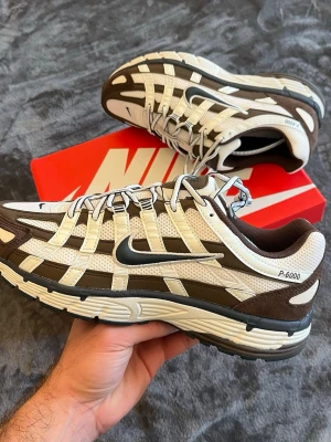 Nike P-6000 sneakers brun/vit - Nike P-6000 sneakers i brunt och vitt med mesh och mockadetaljer. Snygg retrodesign med silvrig swoosh på sidan och coola paneler. Skön sula och låga i modellen, perfekta för en streetwear-look.