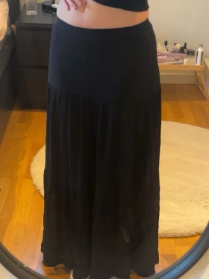 Svart lång maxikjol - Svart maxikjol med hög midja och flowy passform. Kjolen är lång och har en mjuk, lätt rynkad design som ger ett snyggt fall. Perfekt att styla med crop tops eller stickade tröjor. Materialet känns mjukt och bekvämt mot huden.