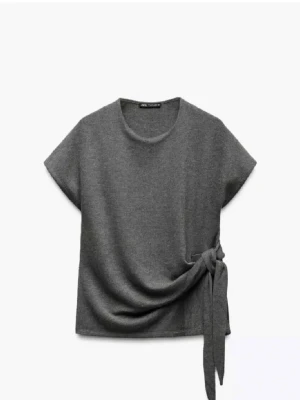 Grå topp med knyt från Zara - Snygg grå topp från Zara med korta ärmar och rund halsringning. Toppen har en cool knytdetalj i sidan som ger en trendig look. Perfekt att styla med jeans eller kjol för en avslappnad men chic outfit.
